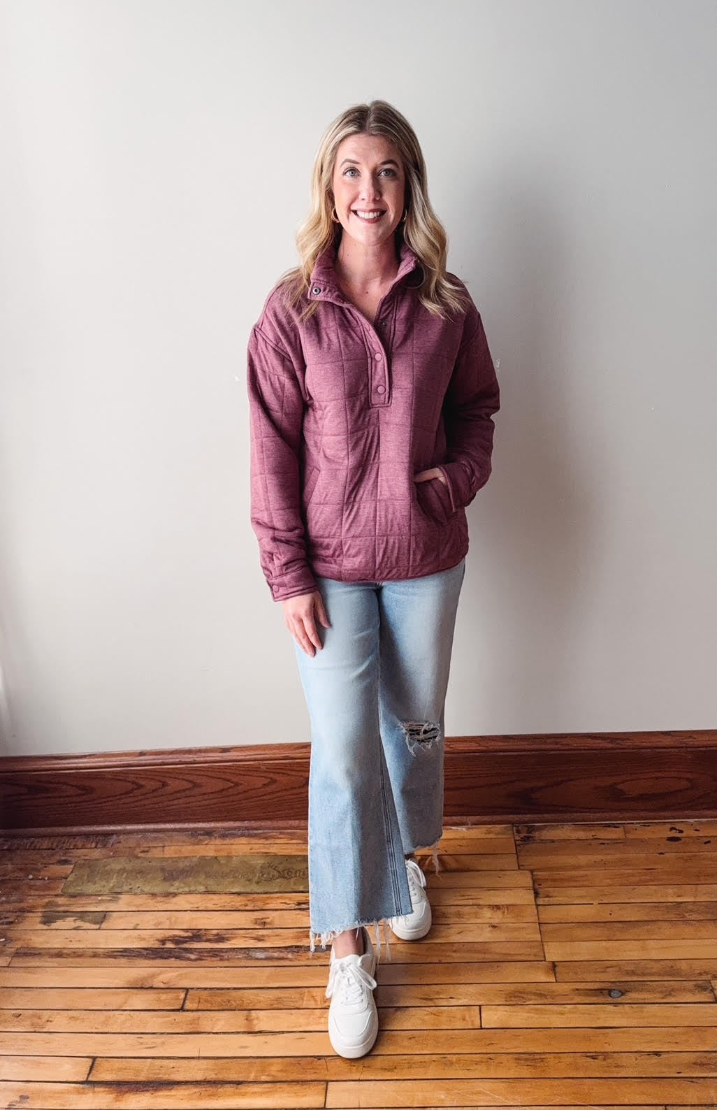 Woman wearing a pink jacket and light blue jeans standing on a wooden floor against a white wall.