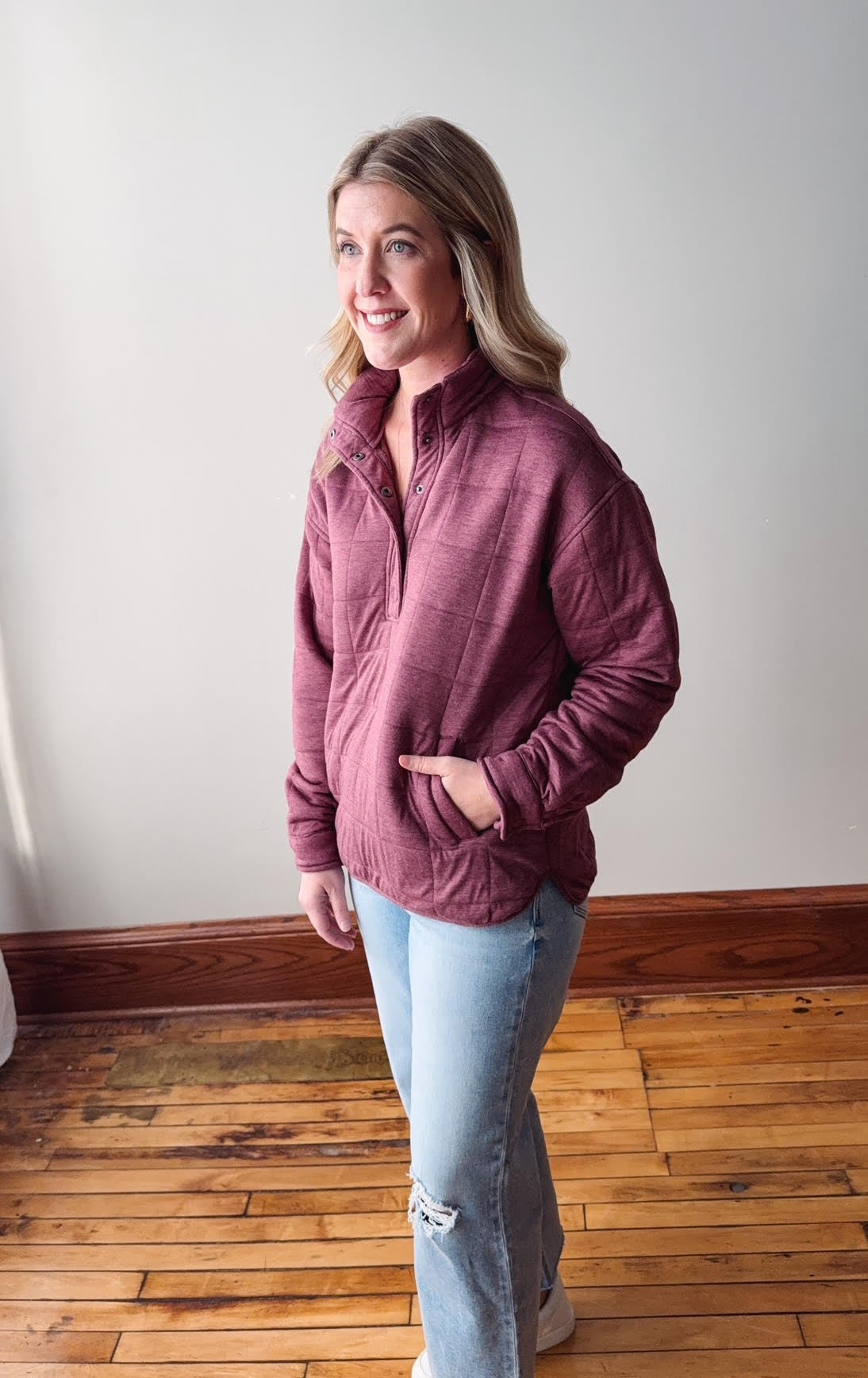 Woman wearing a maroon jacket standing on a wooden floor with a white wall background