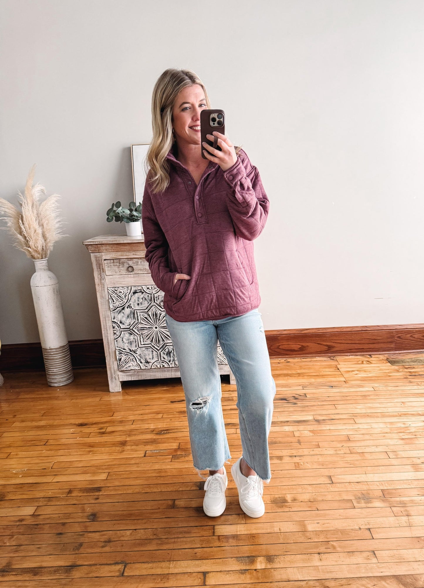 Woman taking a mirror selfie wearing a purple sweater and light blue jeans in a room with wooden flooring.