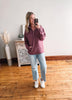 Woman taking a mirror selfie wearing a purple sweater and light blue jeans in a room with wooden flooring.
