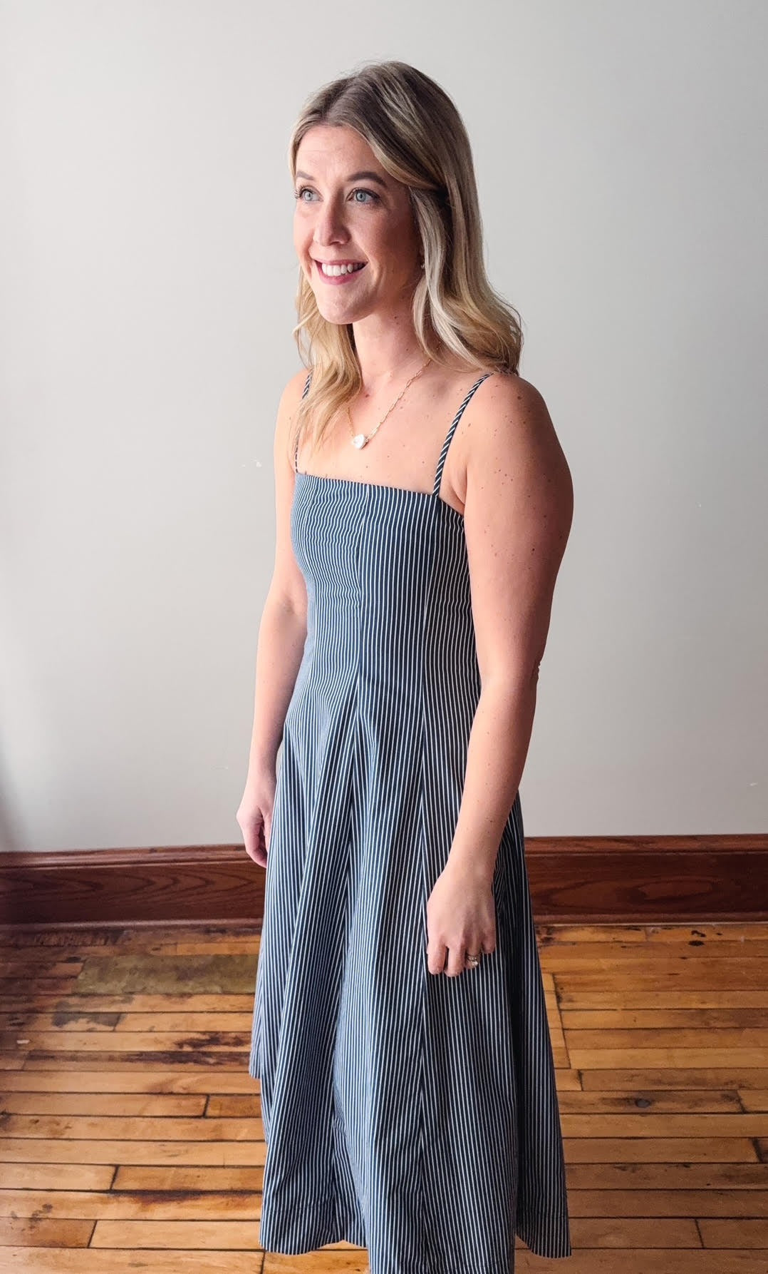 Woman wearing a blue striped dress standing in a room with wooden flooring and a white wall.