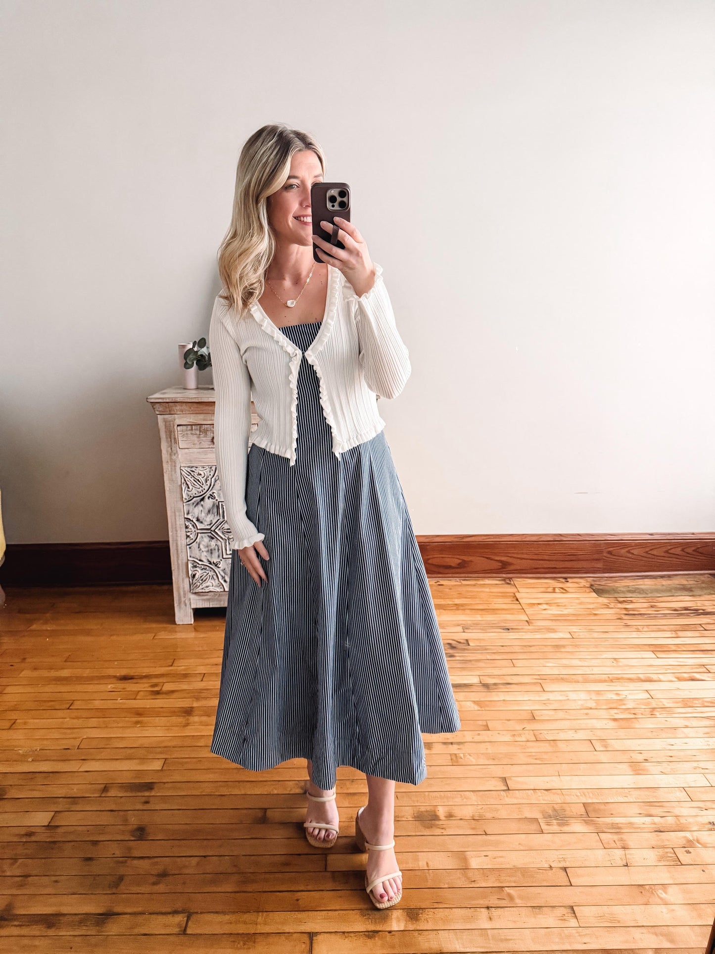 Woman taking a mirror selfie wearing a white cardigan and blue dress on a wooden floor.