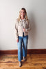 Woman wearing a beige jacket and blue jeans standing against a plain wall.