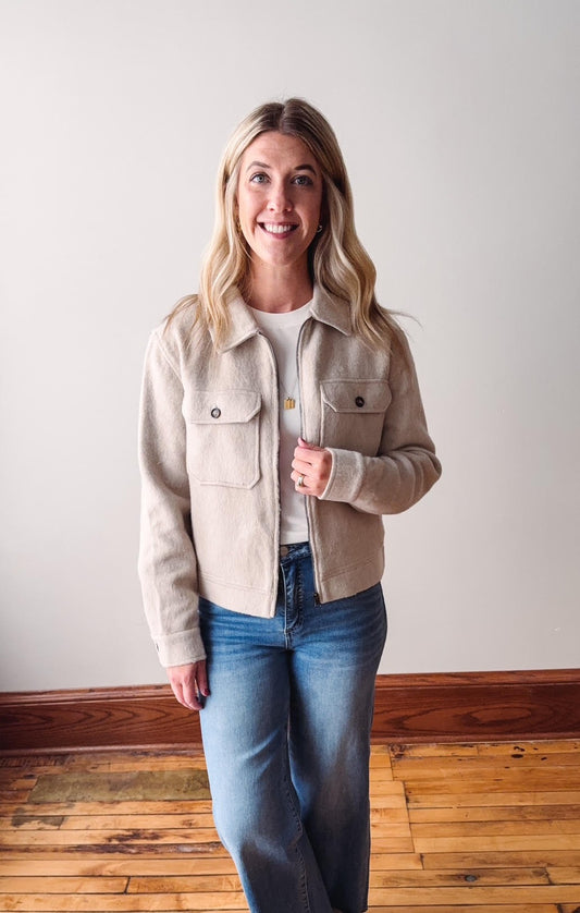 Woman wearing a beige jacket and blue jeans standing against a white wall.