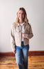 Woman wearing a beige jacket and blue jeans standing against a white wall.