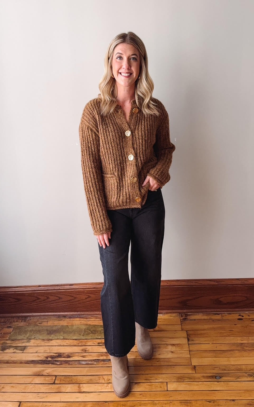 Woman wearing a brown knitted cardigan and dark pants standing on a wooden floor.