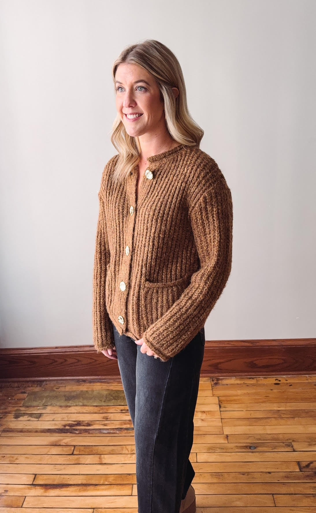 Woman wearing a brown knitted cardigan standing on a wooden floor with a white wall background