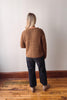Person wearing a brown sweater and dark pants standing against a white wall on a wooden floor.