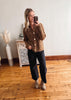 Woman taking a mirror selfie wearing a brown knitted cardigan, black pants, and beige shoes in a room with wooden flooring and a decorative cabinet.