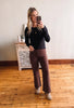 Woman taking a mirror selfie wearing a black top and brown pants in a room with wooden flooring.