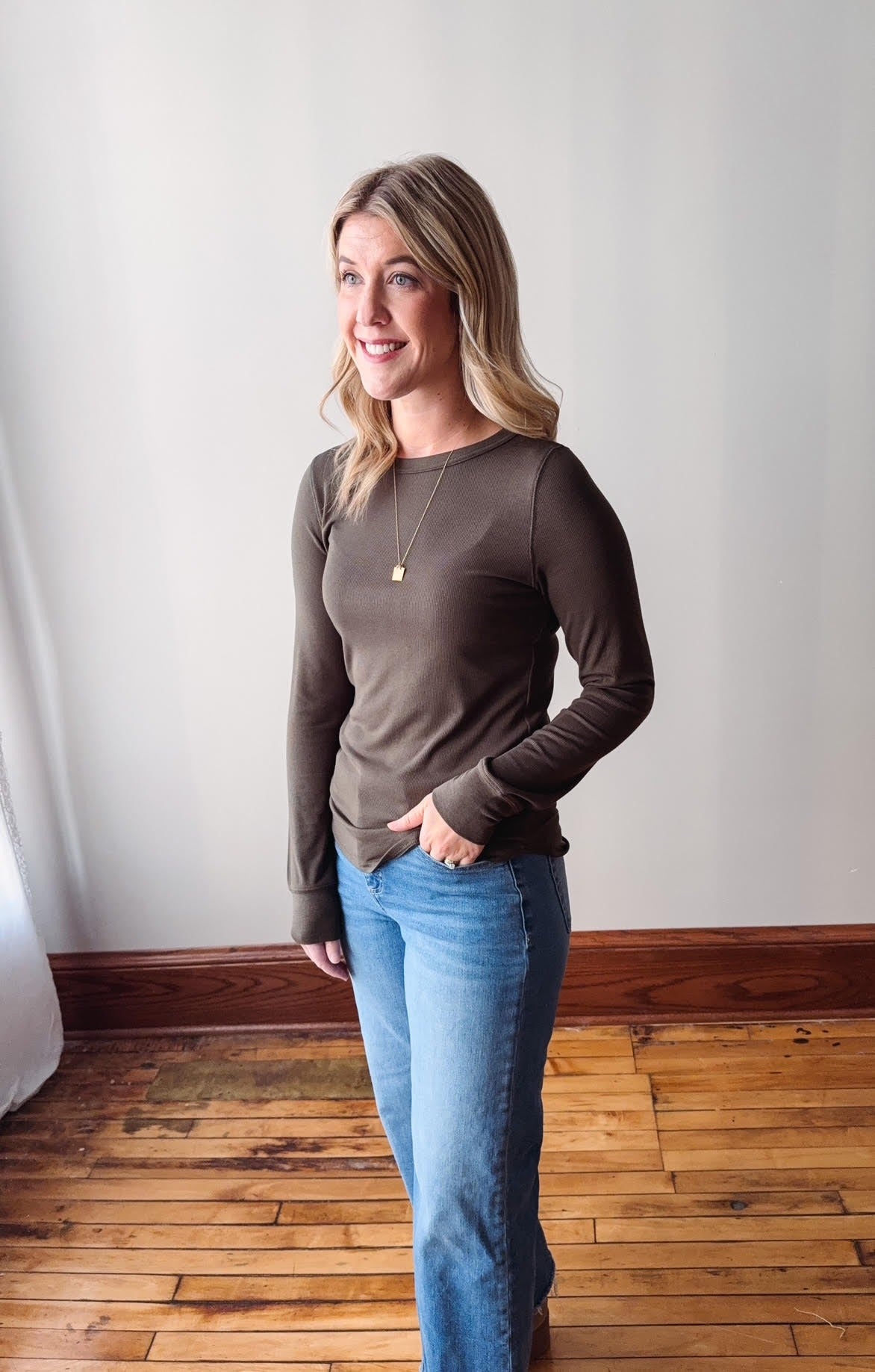 Woman wearing a brown long-sleeve top and blue jeans standing in a room with wooden flooring and a white wall.