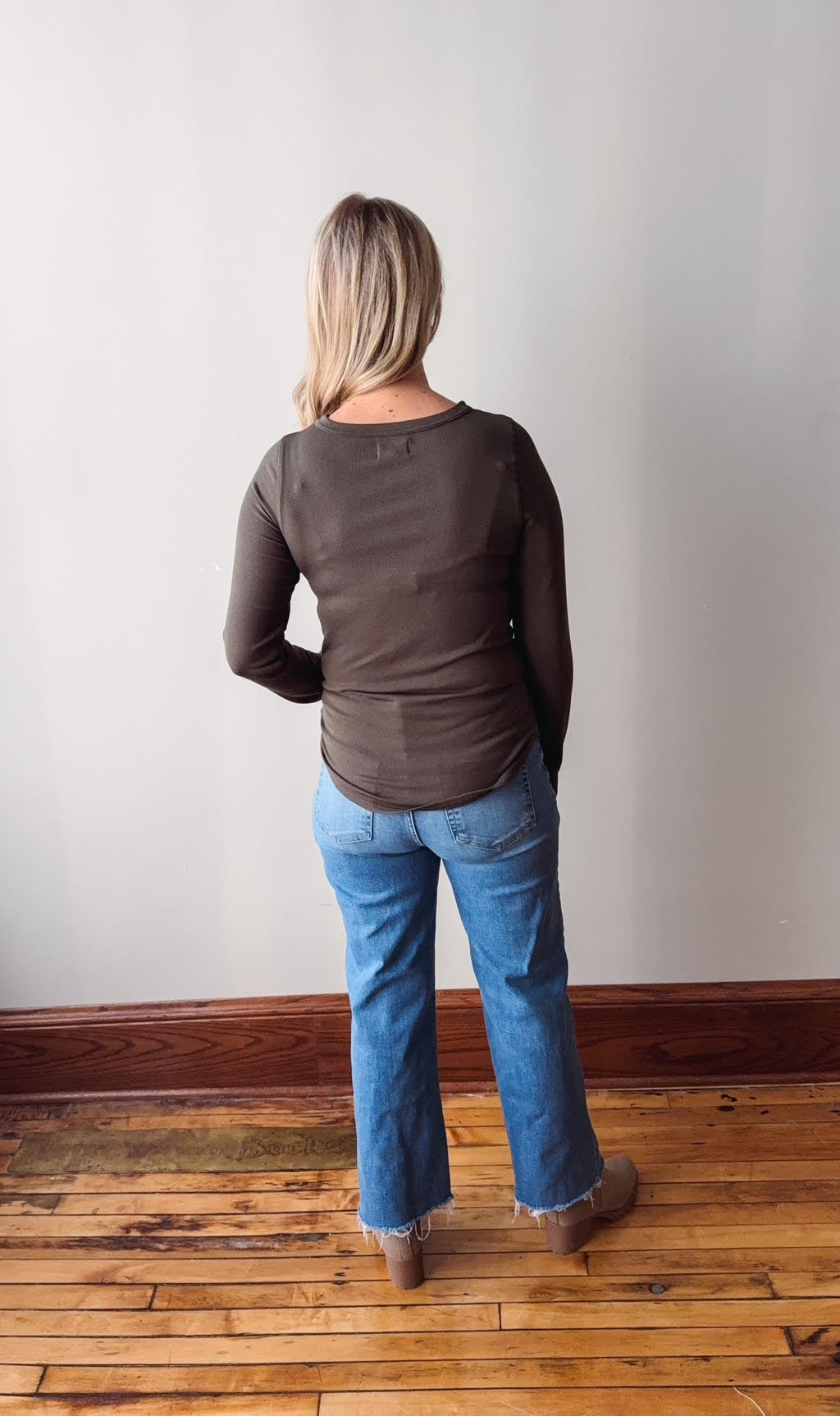 Person wearing a brown long-sleeve shirt and blue jeans standing against a plain wall.