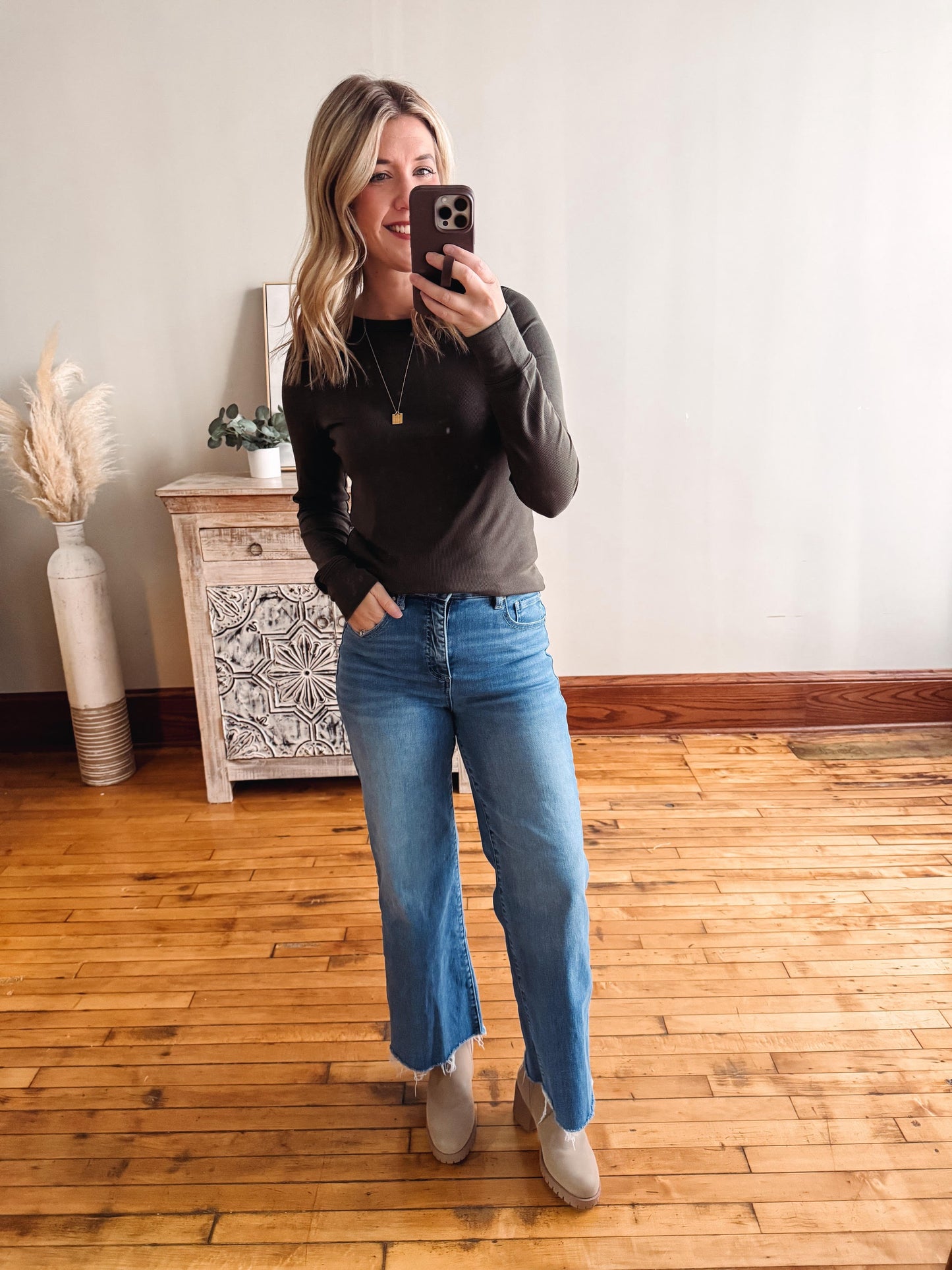 Woman taking a mirror selfie wearing a dark sweater and blue jeans in a room with wooden flooring.