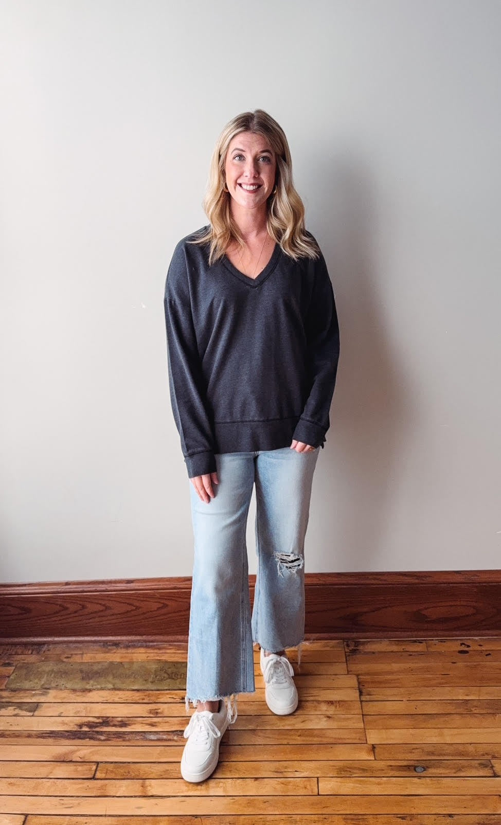 Woman wearing a dark blue sweater and light blue jeans standing on a wooden floor with a white wall background.