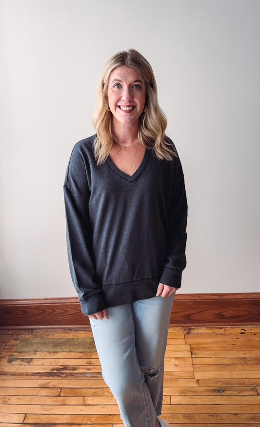 Woman wearing a dark gray sweater and light blue jeans standing on a wooden floor with a white wall background.