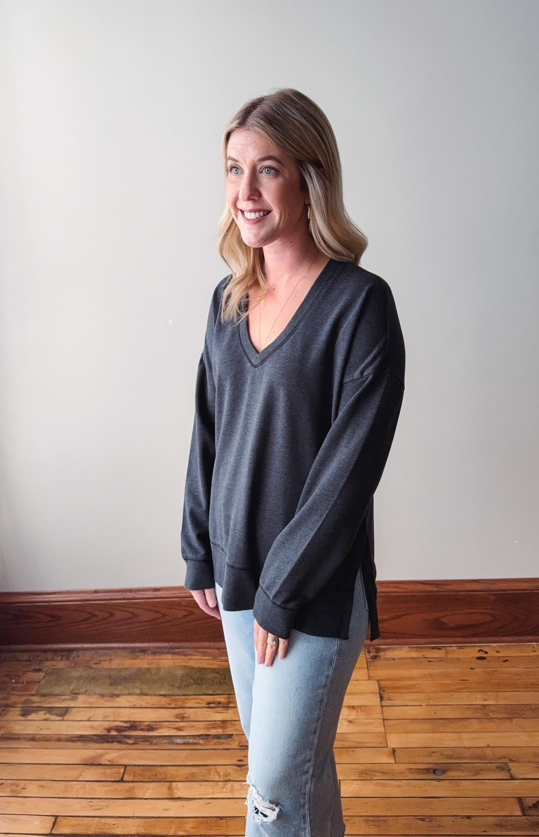 Woman wearing a dark gray sweater and light blue jeans standing on a wooden floor with a white wall background.