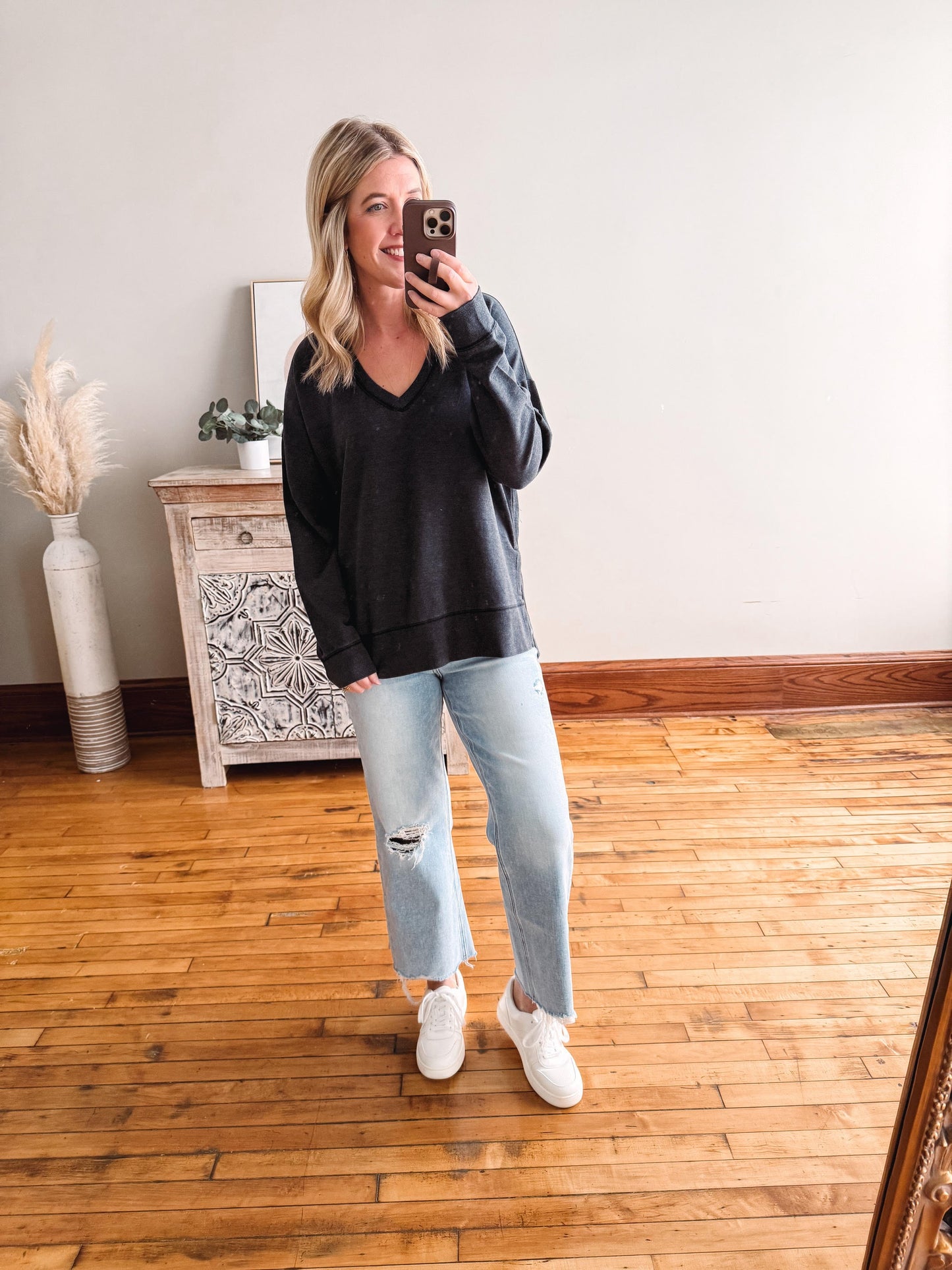 Woman taking a mirror selfie wearing a black sweater and light blue jeans in a room with wooden flooring.