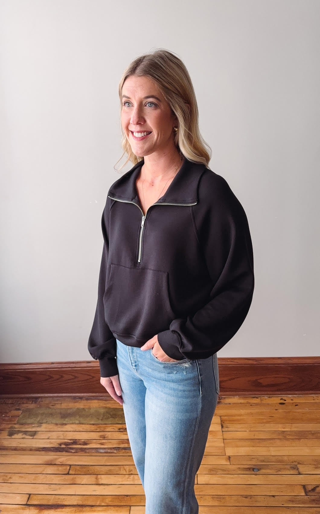 Woman wearing a black pullover with a zipper and light blue jeans against a plain wall.