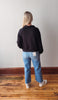 Person wearing a black sweater and blue jeans standing on a wooden floor with a white wall background