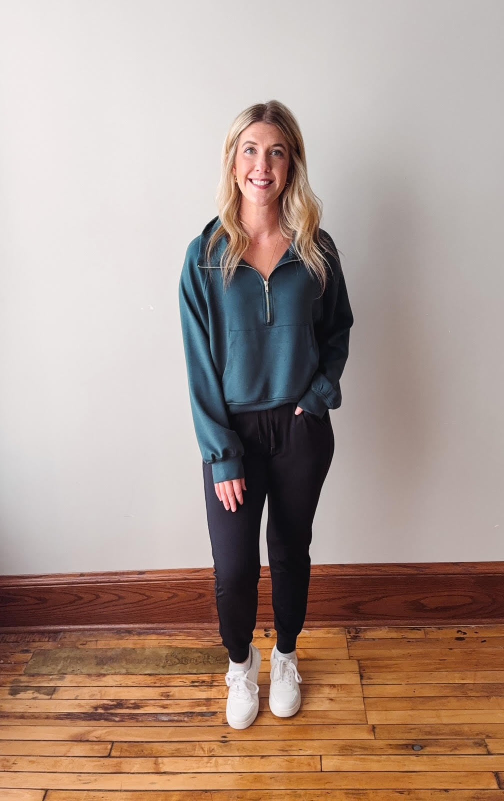 Woman wearing a teal sweatshirt and black pants standing on a wooden floor with a white wall background