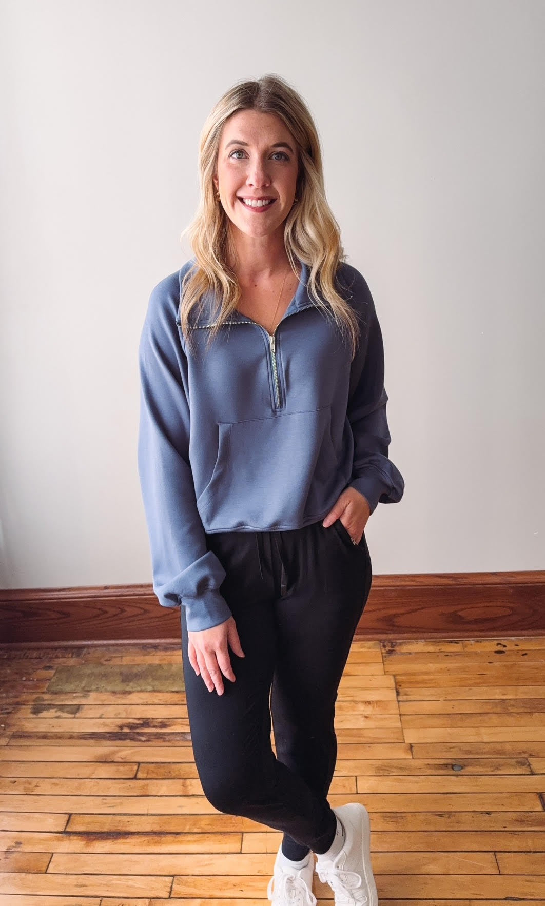 Woman wearing a blue pullover and black leggings standing on a wooden floor.