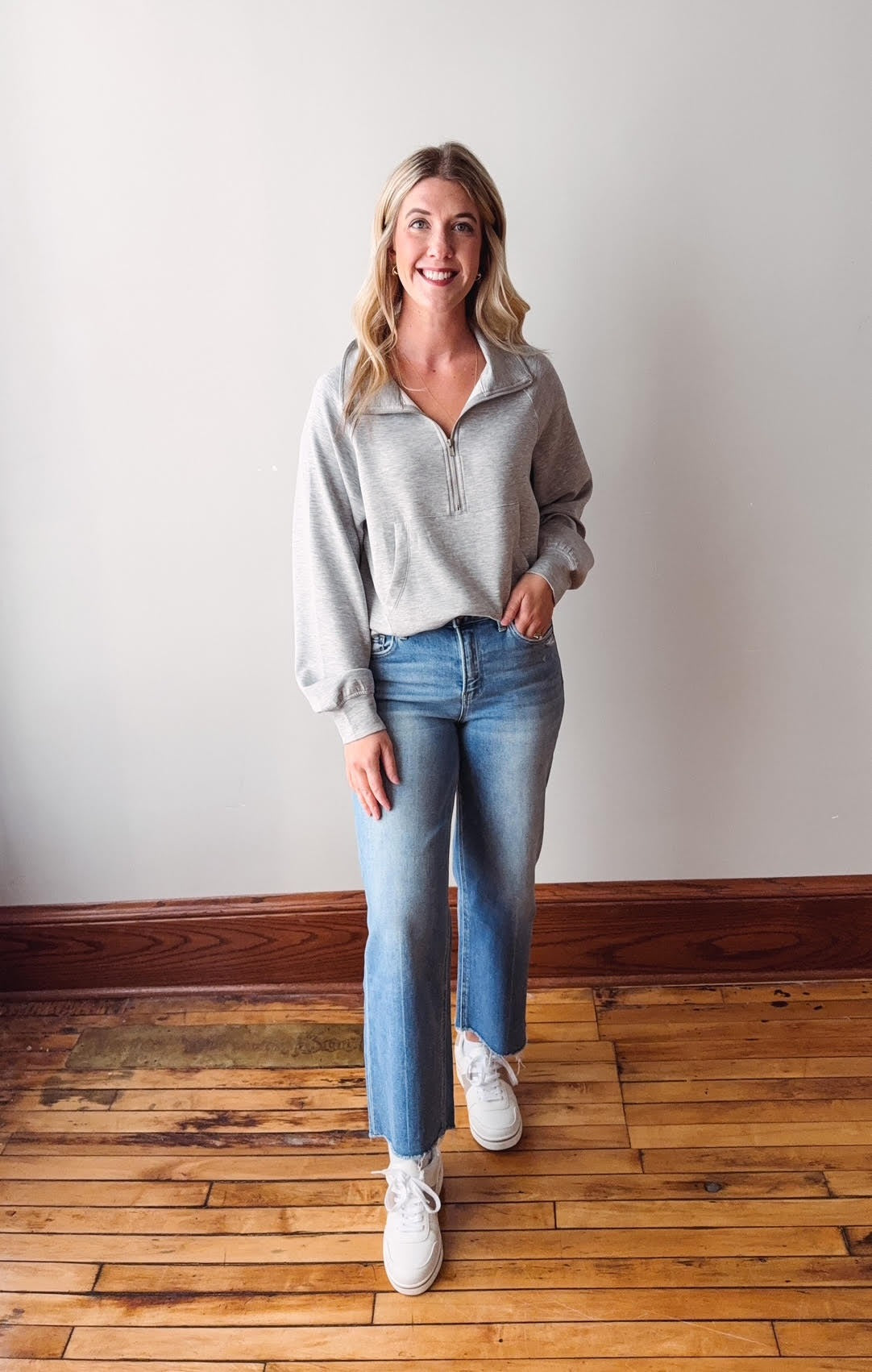 Woman wearing a light gray sweater and blue jeans standing on a wooden floor.