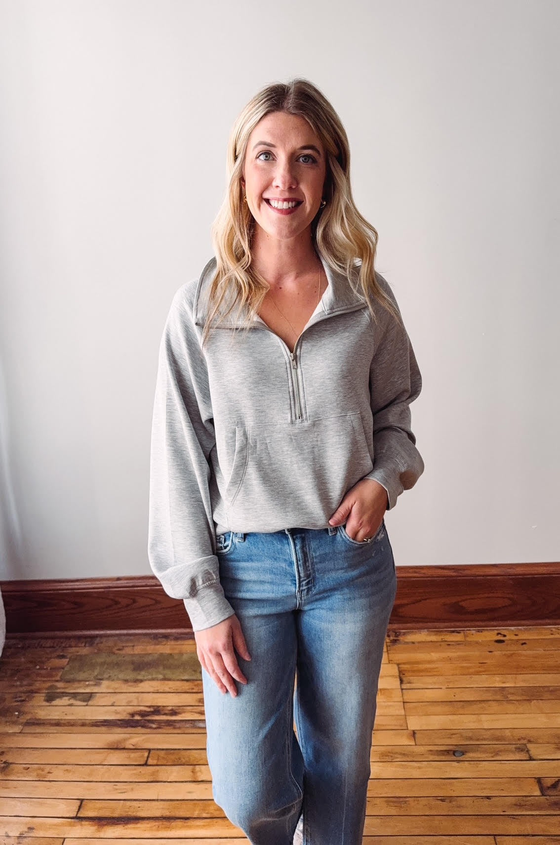 Woman wearing a gray long-sleeve top and blue jeans standing against a white wall.