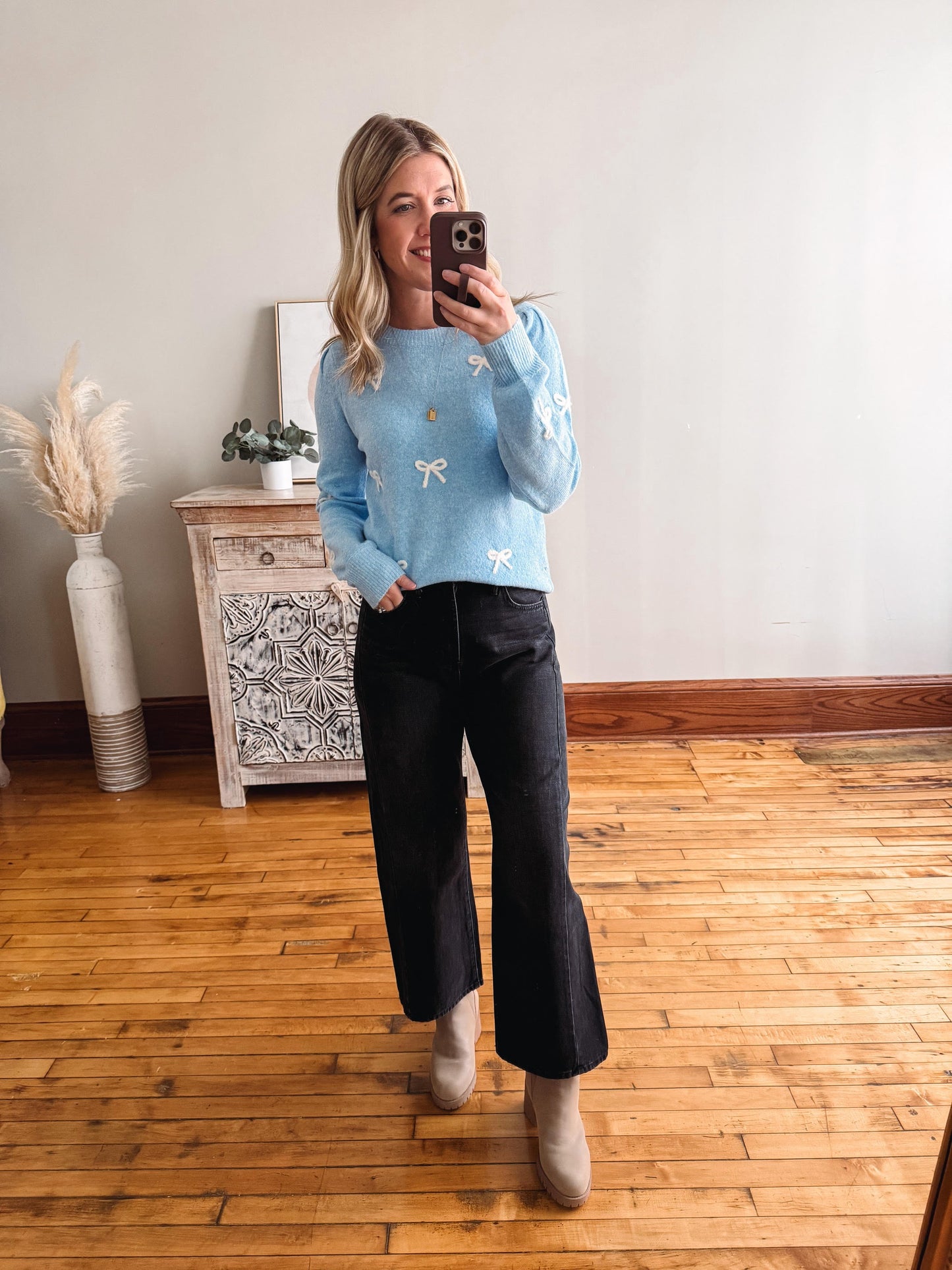 Person wearing a light blue sweater with white patterns and black pants, standing in a room with wooden flooring and a decorative cabinet.