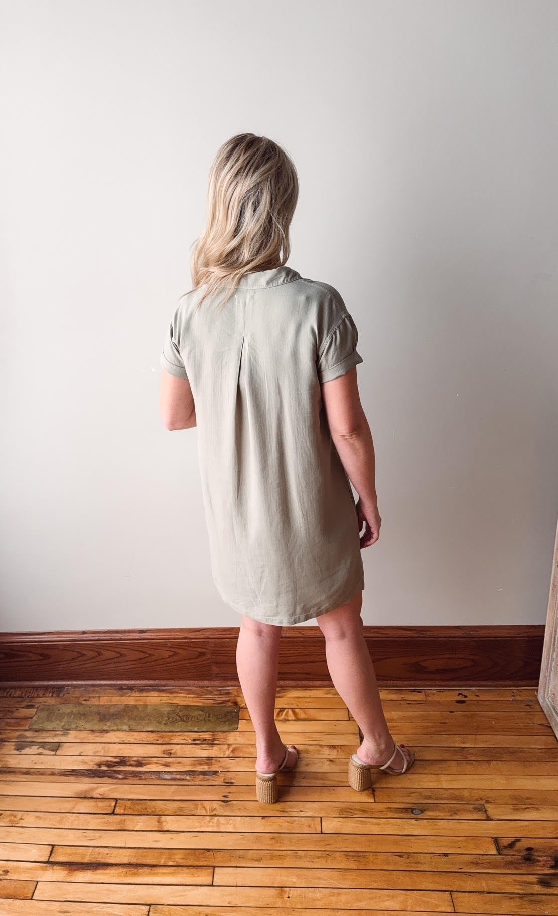 Woman wearing a light green dress standing on a wooden floor against a white wall.