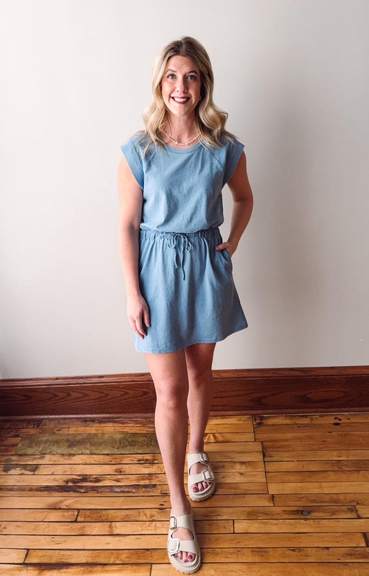 Woman wearing a light blue dress standing on a wooden floor with a white wall background