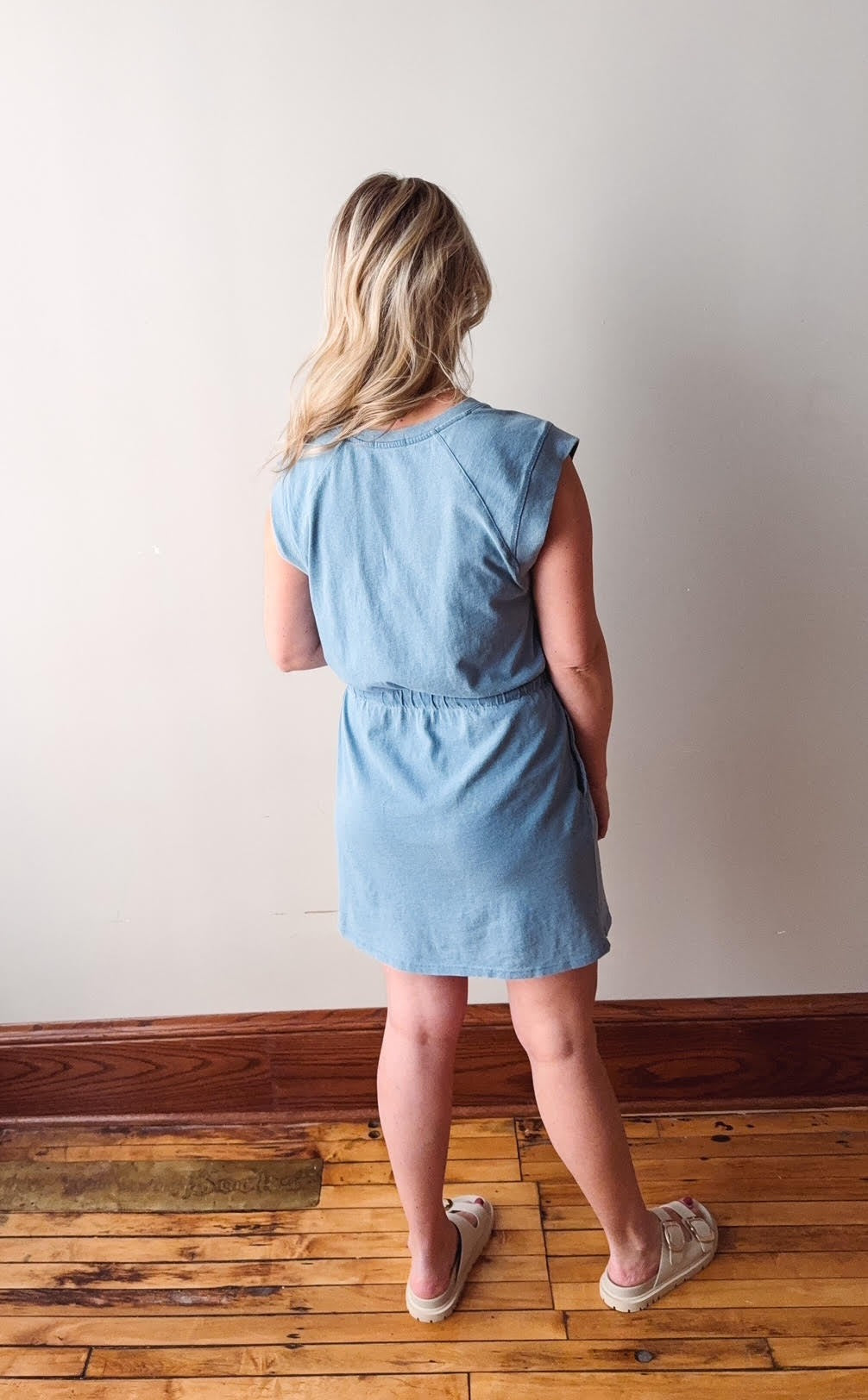 Woman wearing a light blue dress standing on a wooden floor against a white wall.