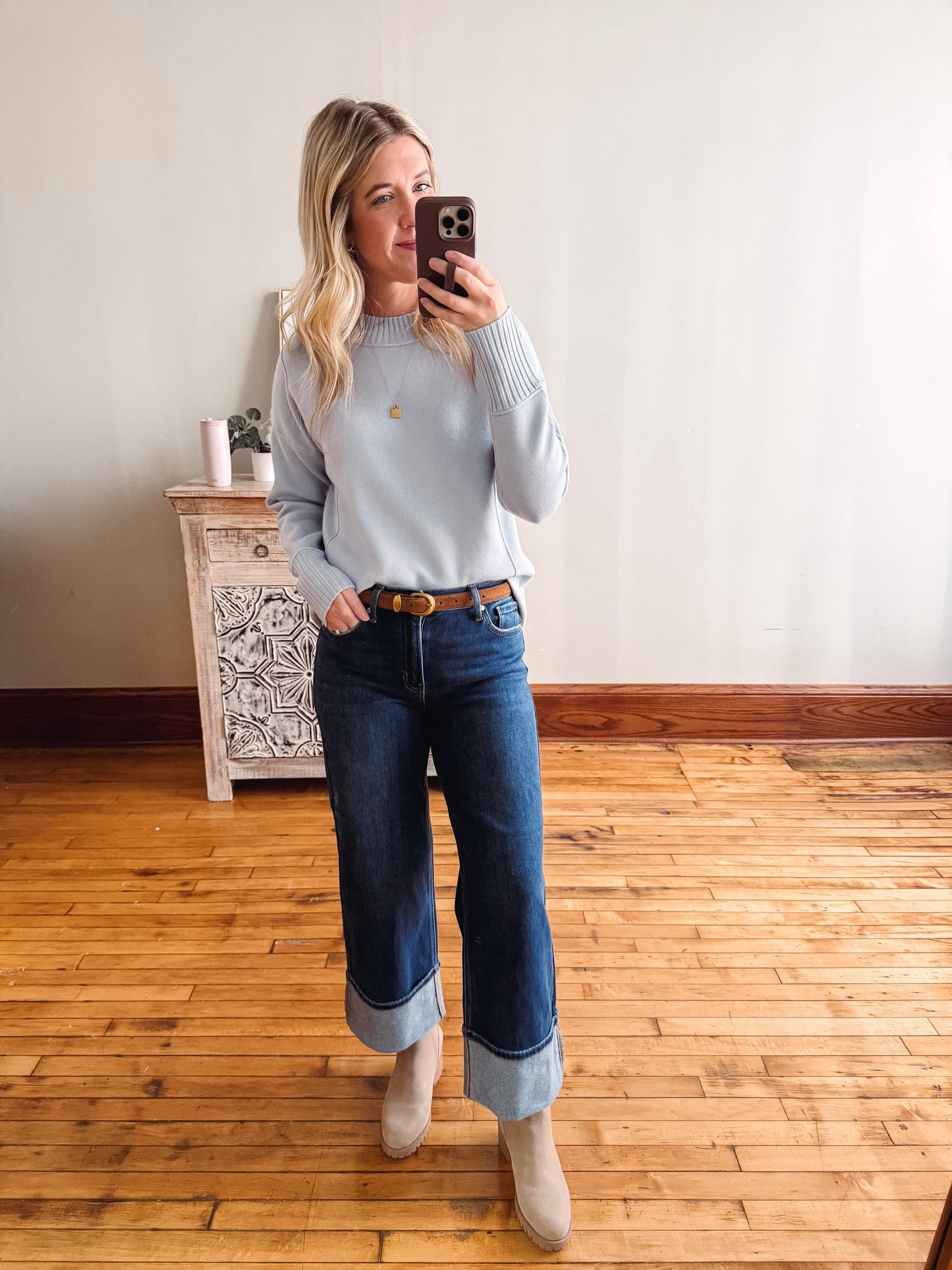 Woman taking a mirror selfie wearing a light blue sweater and blue jeans in a room with wooden flooring.