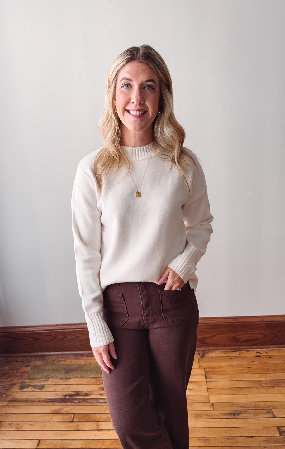 Woman wearing a white sweater and brown pants standing on a wooden floor with a white wall background