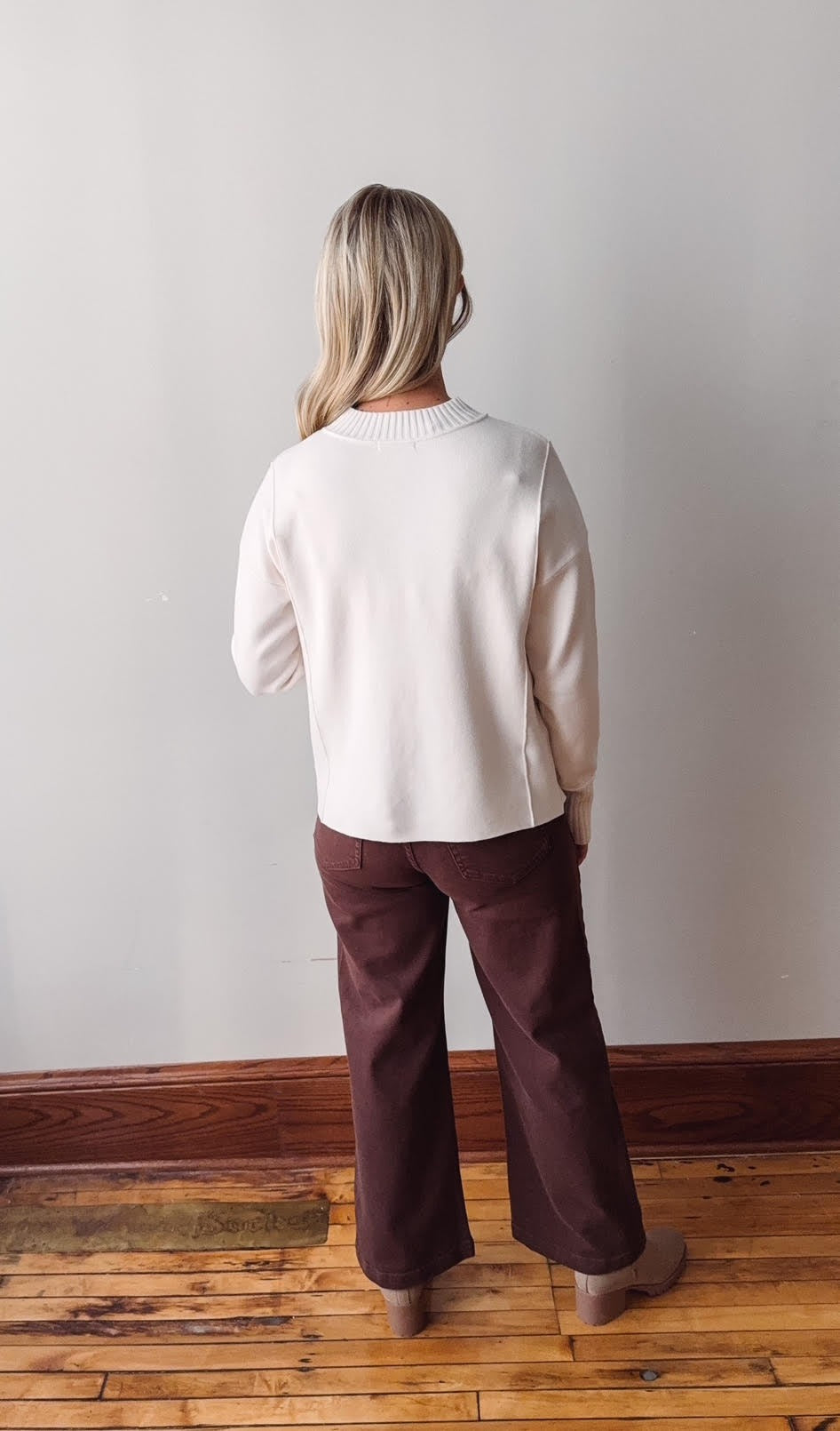 Person wearing a white sweater and brown pants standing against a plain wall.