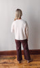 Person wearing a white sweater and brown pants standing against a plain wall.
