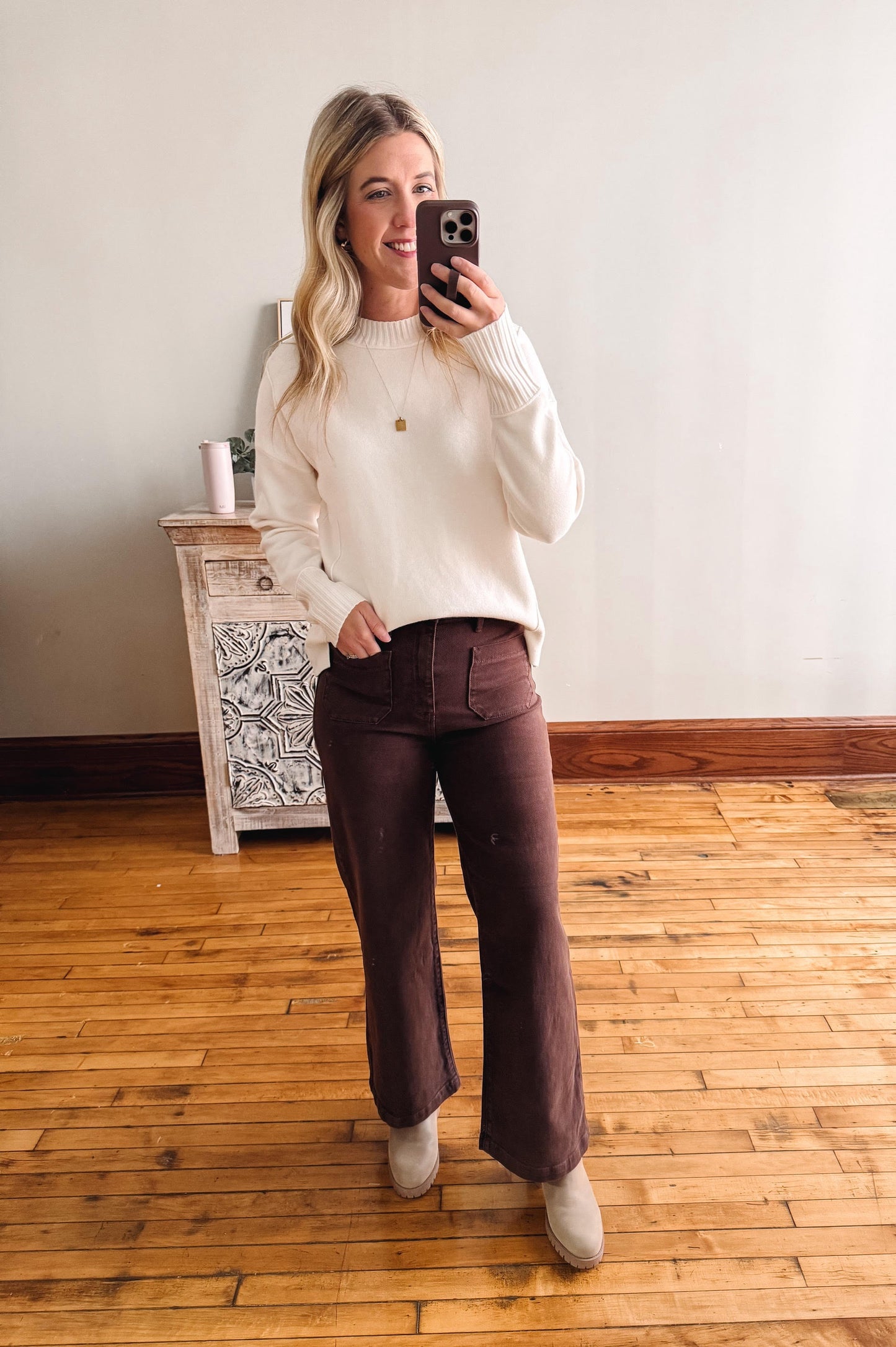 Woman taking a mirror selfie wearing a white sweater and brown pants in a room with wooden flooring.