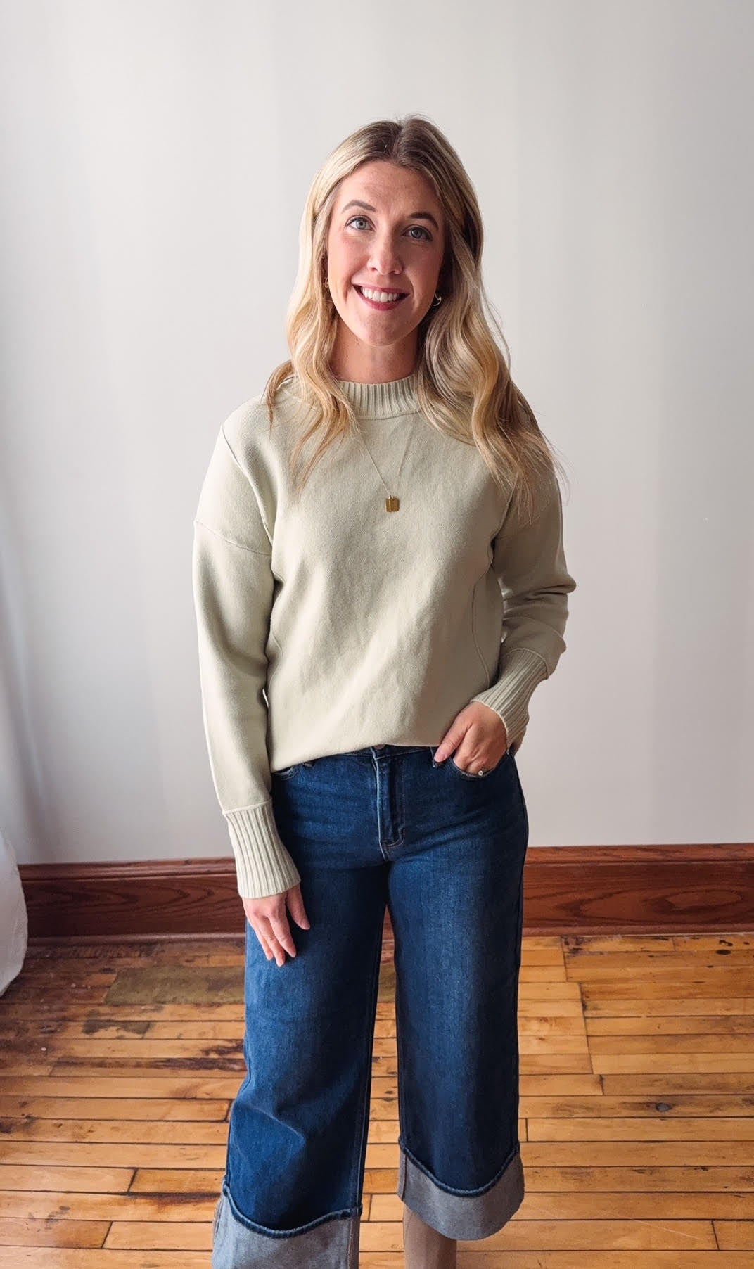 Woman wearing a sage sweater and blue jeans standing in a room with wooden flooring and a white wall.