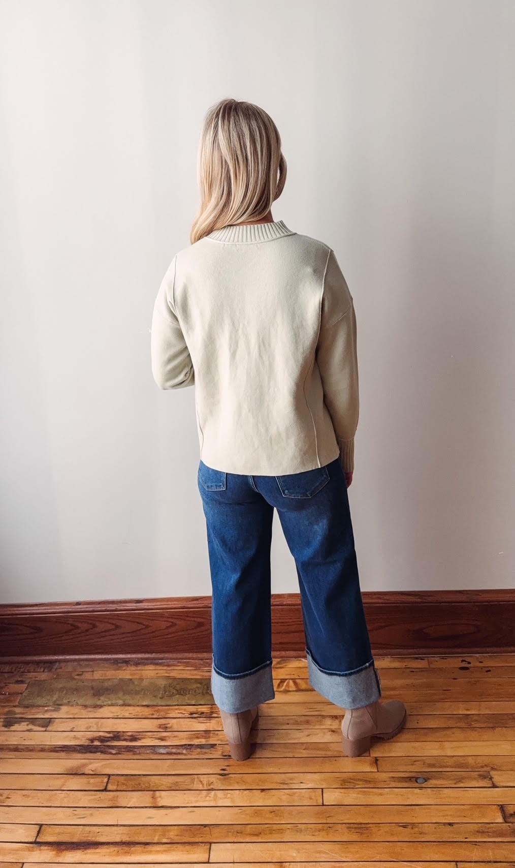 Person wearing a sage sweater and blue jeans standing in a room with wooden flooring and a white wall.