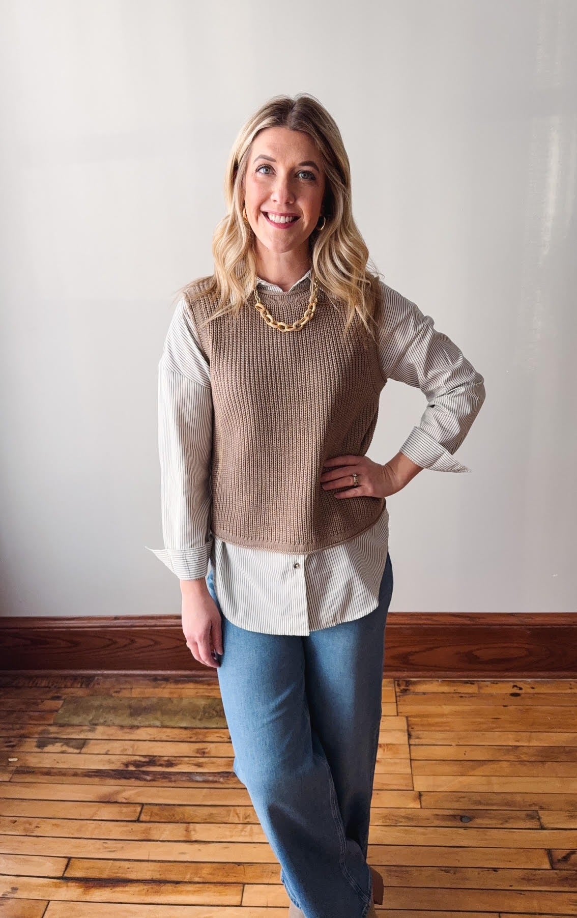 Woman wearing a beige vest over a white shirt with blue pants on a wooden floor.