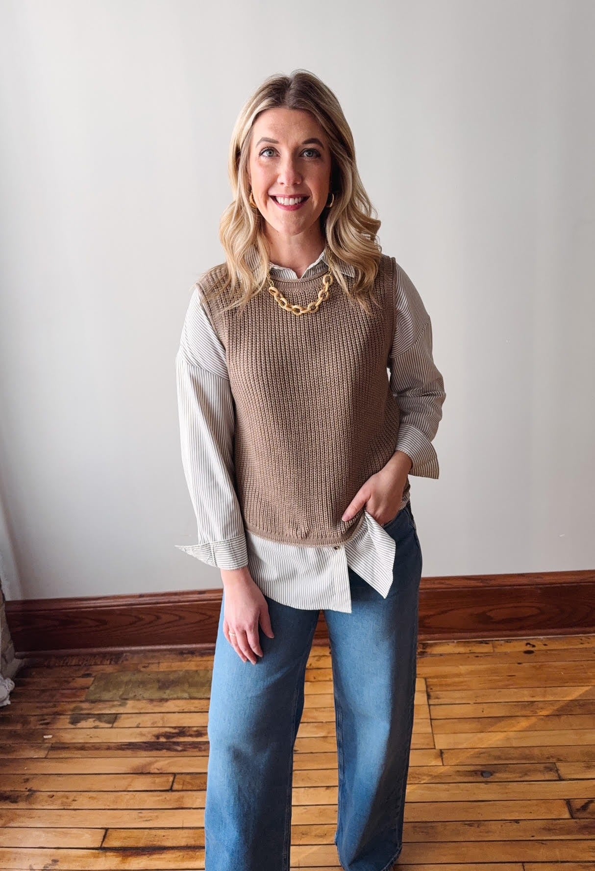 Woman wearing a beige sweater vest over a light shirt and blue jeans, standing in a room with wooden flooring and a white wall.