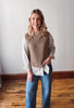 Woman wearing a beige sweater vest over a light shirt and blue jeans, standing in a room with wooden flooring and a white wall.
