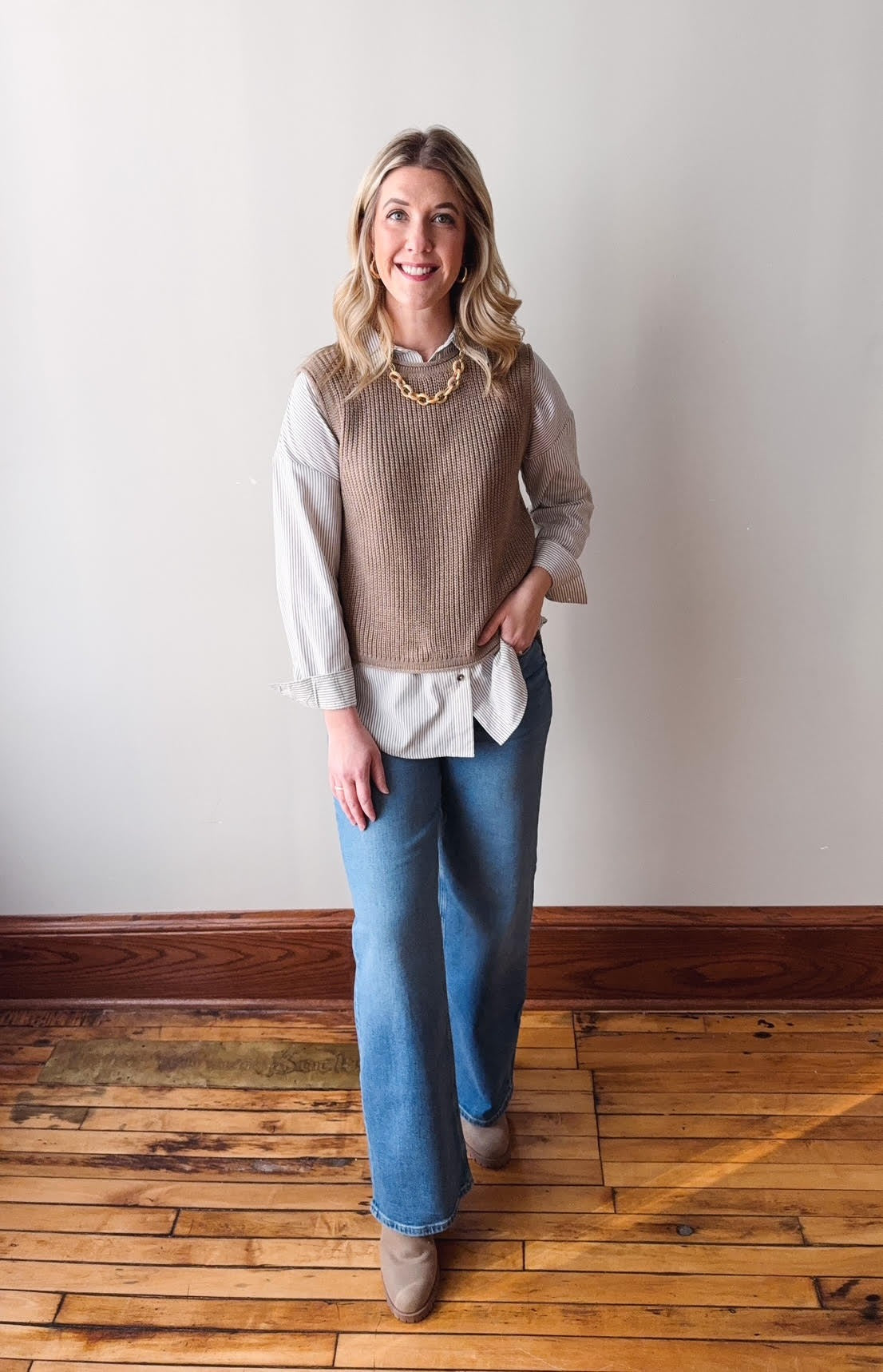 Woman wearing a beige vest over a white shirt with blue jeans on a wooden floor.