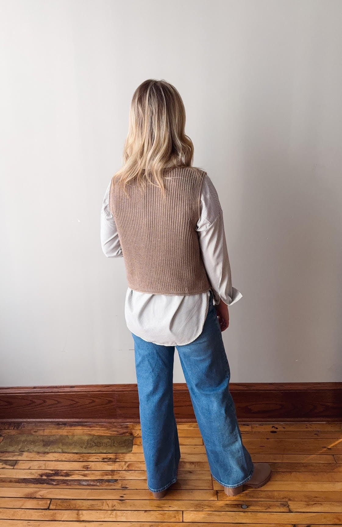 Person wearing a brown vest over a white shirt and blue jeans, standing against a plain wall.