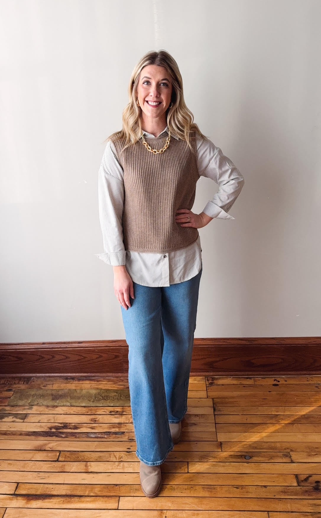Woman wearing a brown sweater vest over a white shirt with blue jeans on a wooden floor.