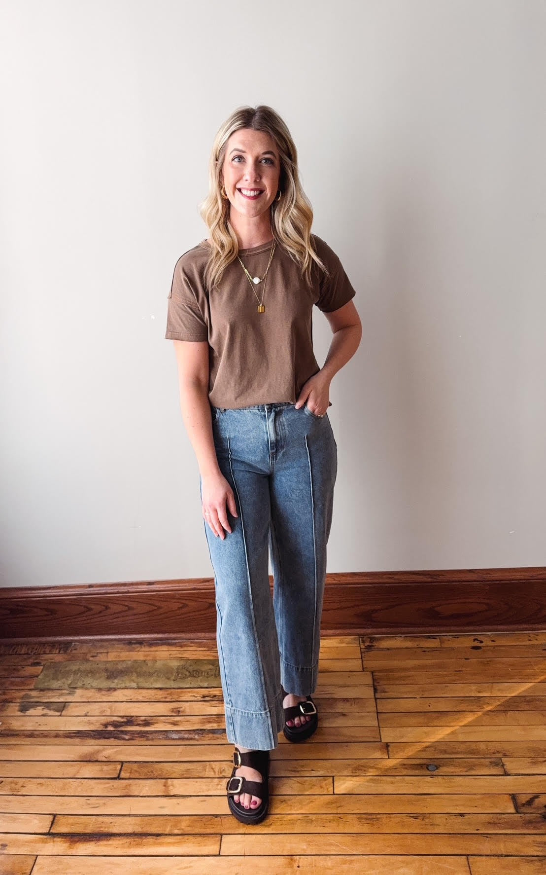 Woman wearing a brown t-shirt and blue jeans standing on a wooden floor.
