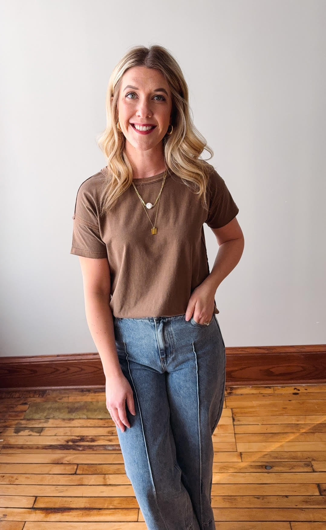 Woman wearing a brown t-shirt and blue jeans standing on a wooden floor.