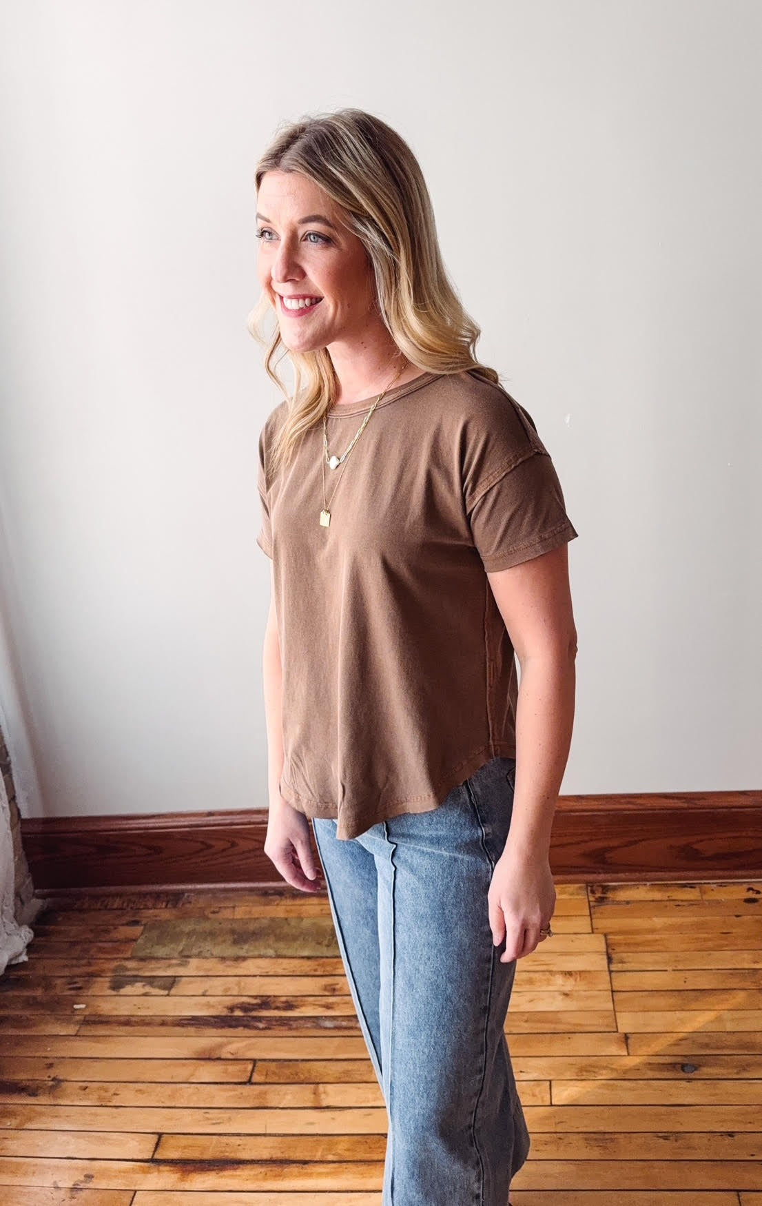 Woman wearing a brown top and blue jeans standing in a room with wooden flooring.