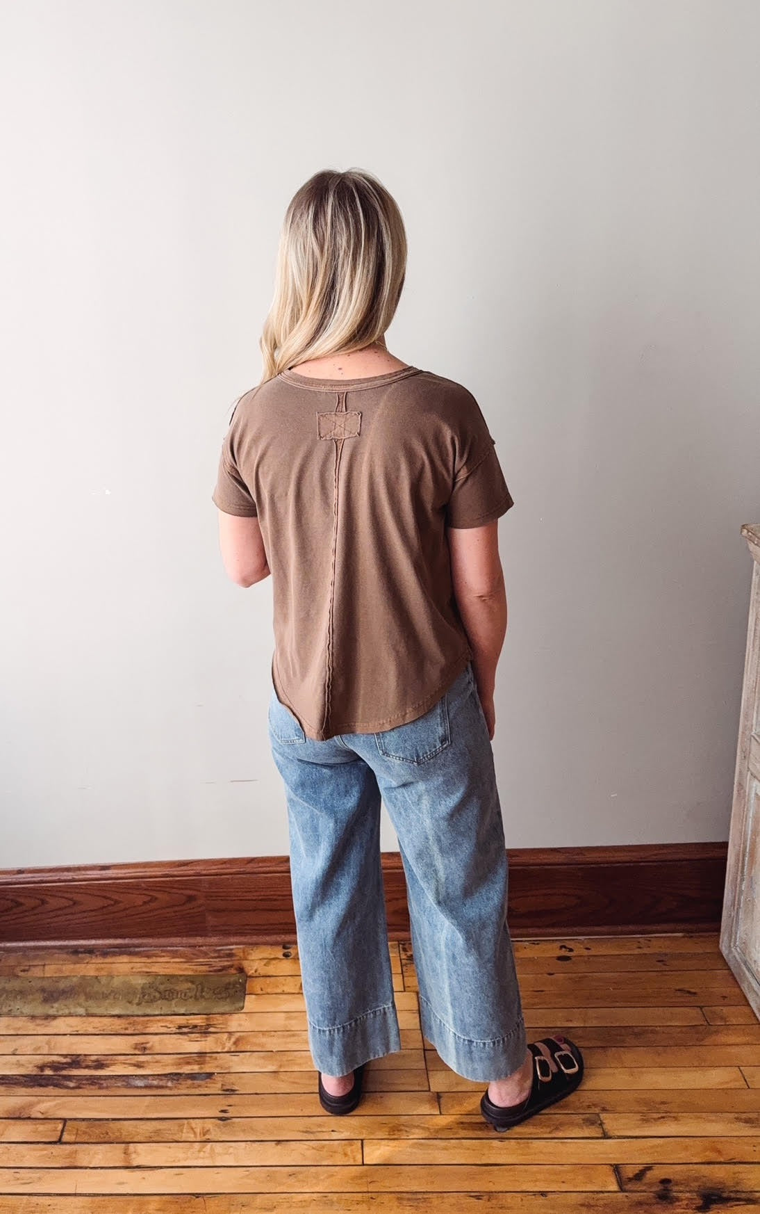 Person wearing a brown shirt and blue jeans standing on a wooden floor against a white wall.
