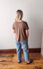 Person wearing a brown shirt and blue jeans standing on a wooden floor against a white wall.