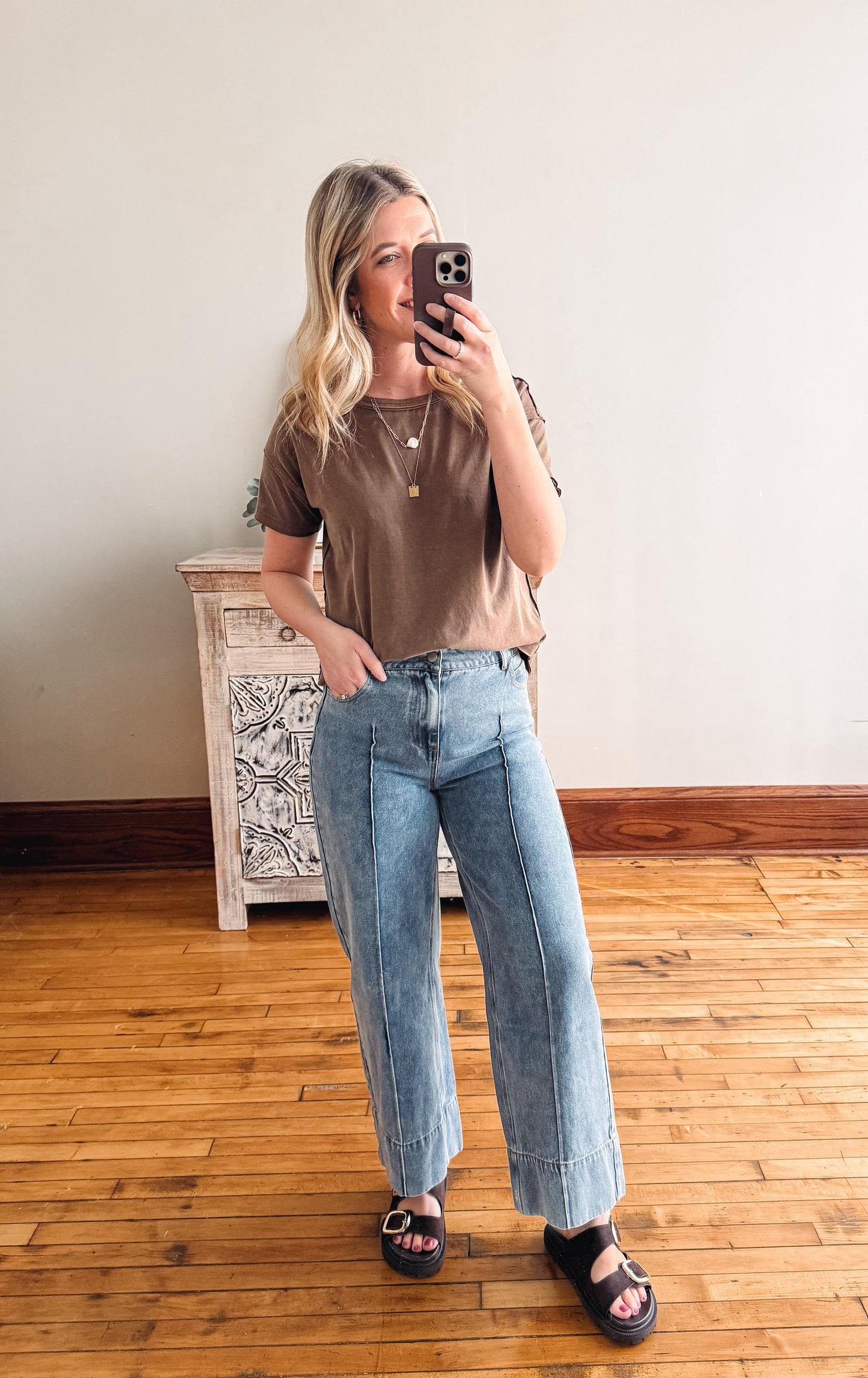 Woman taking a mirror selfie wearing a brown top and blue jeans in a room with wooden flooring.